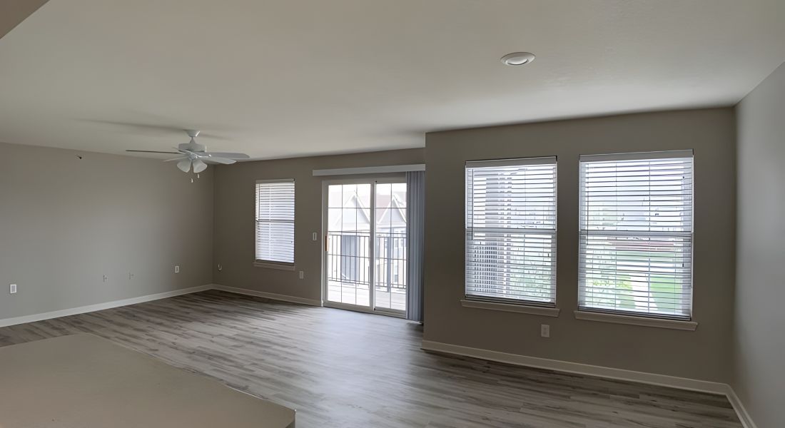 Spacious, empty living room with gray walls, wood floors, large windows, and a ceiling fan.