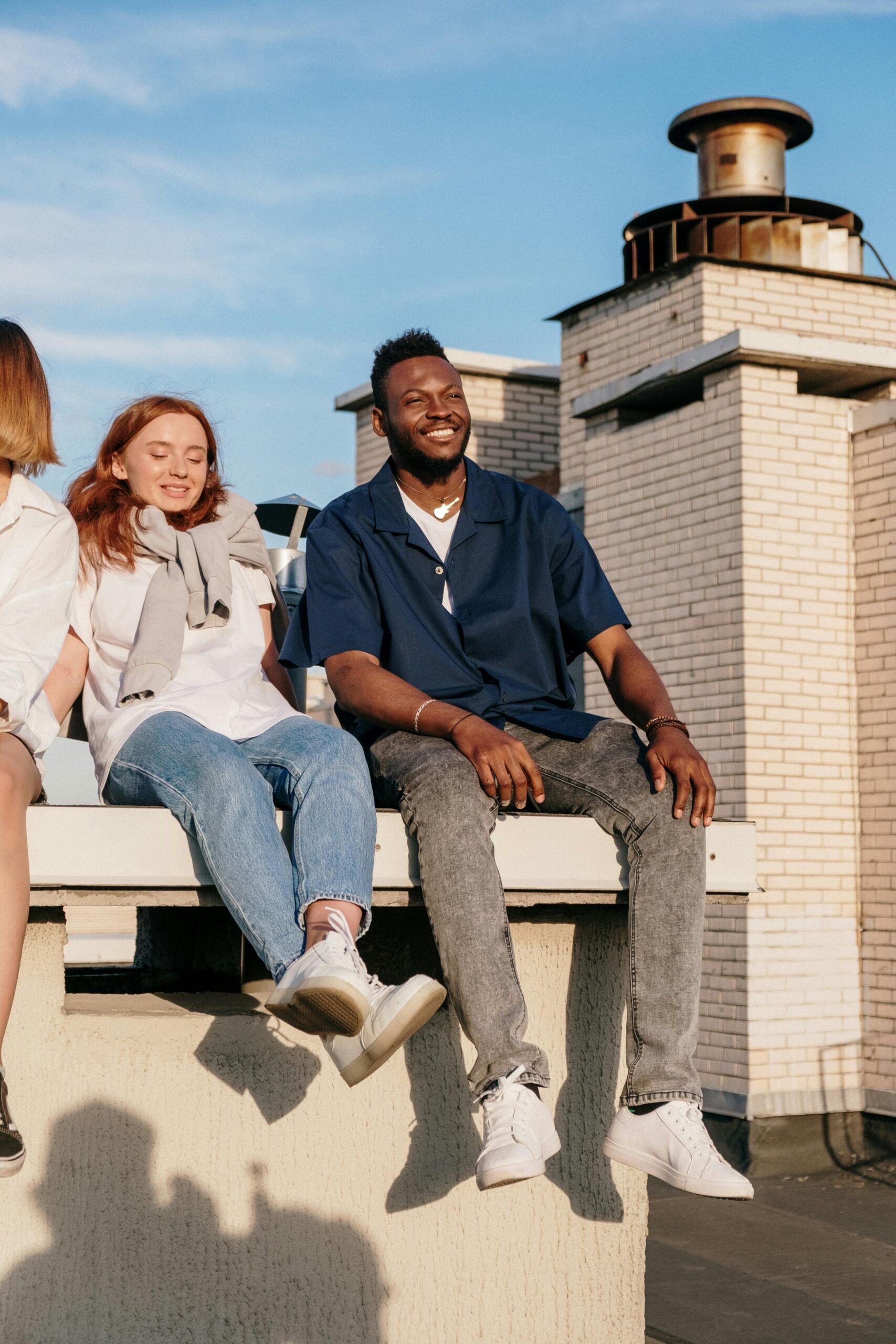 Two friends sit on a rooftop ledge, smiling and enjoying the sunny weather next to a brick chimney.