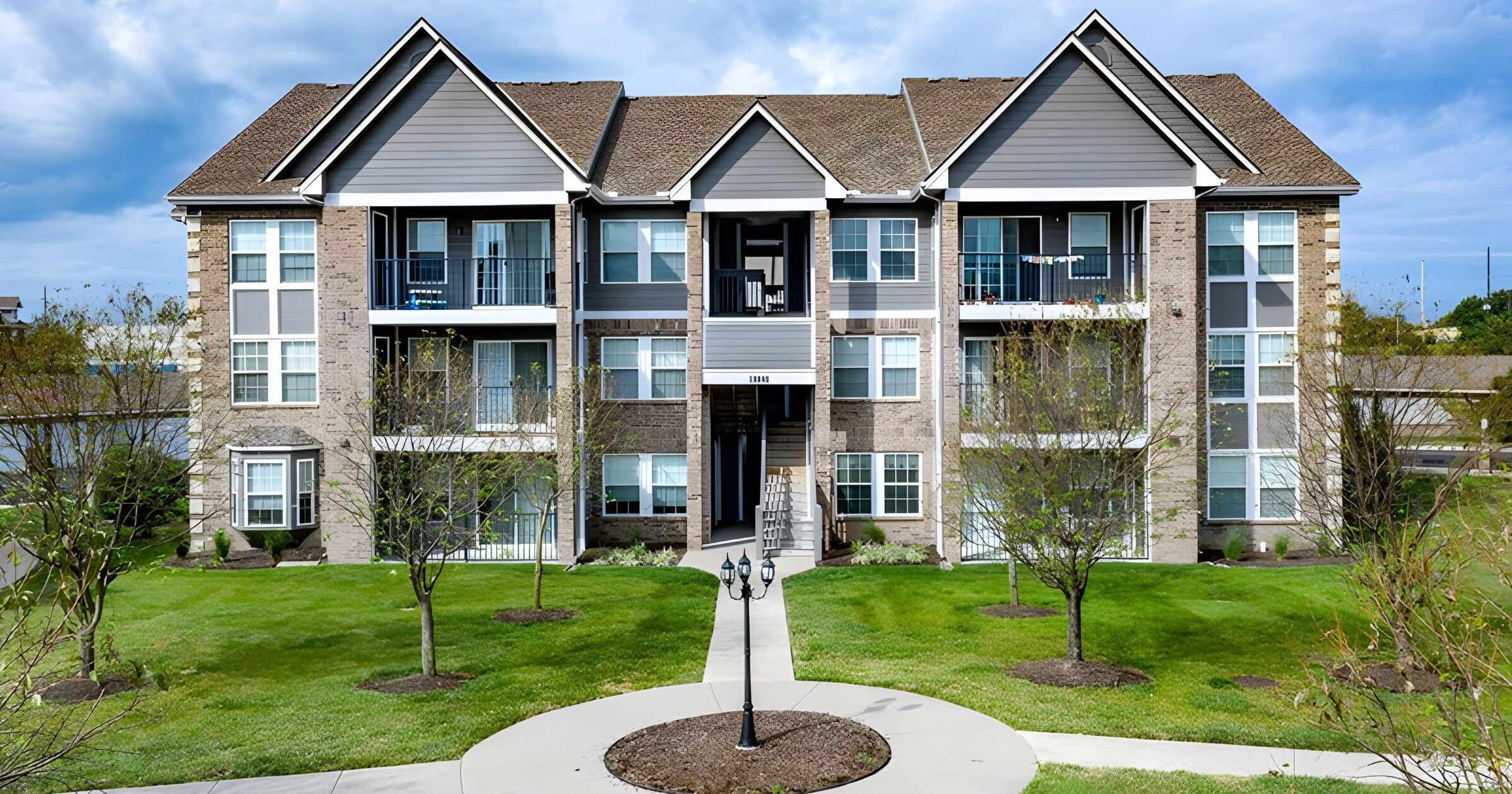 Three-story apartment building with balconies, landscaped lawn, and a sidewalk leading to the main entrance.