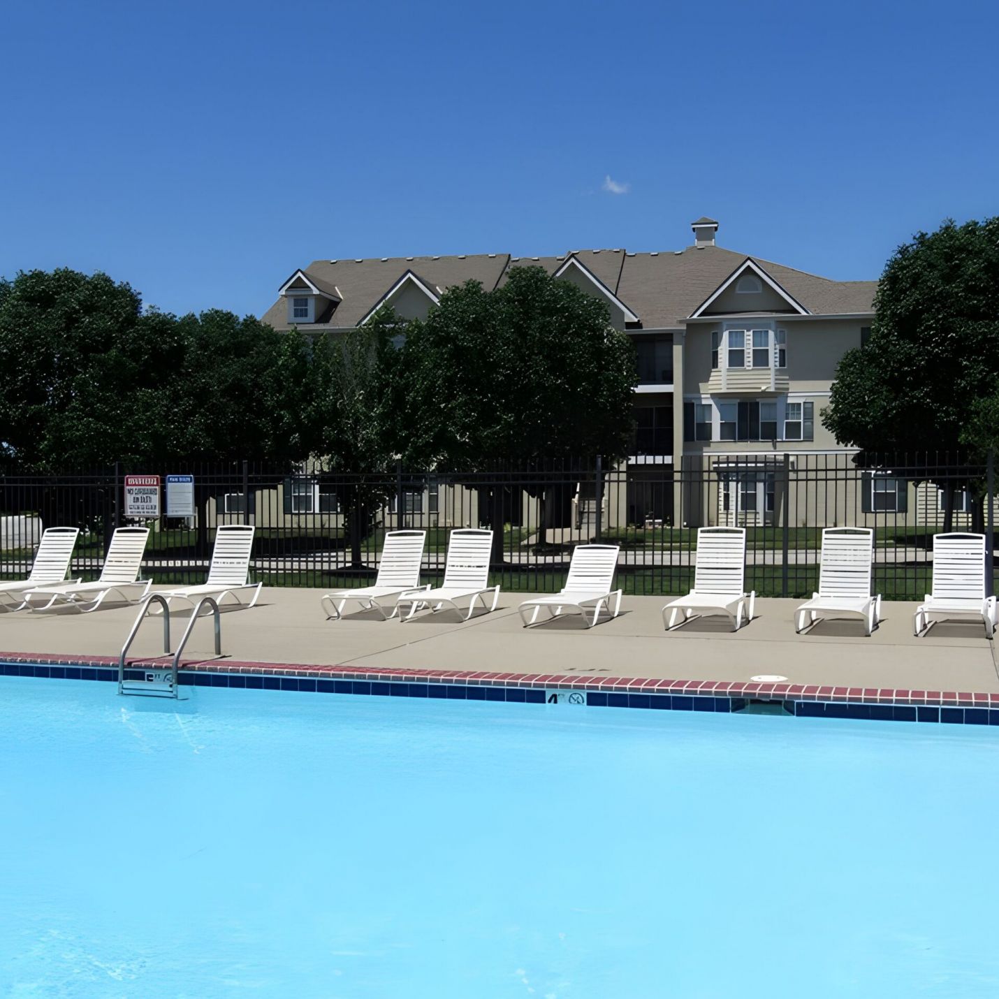 Outdoor swimming pool with empty white lounge chairs, a fence, and an apartment building in the background.