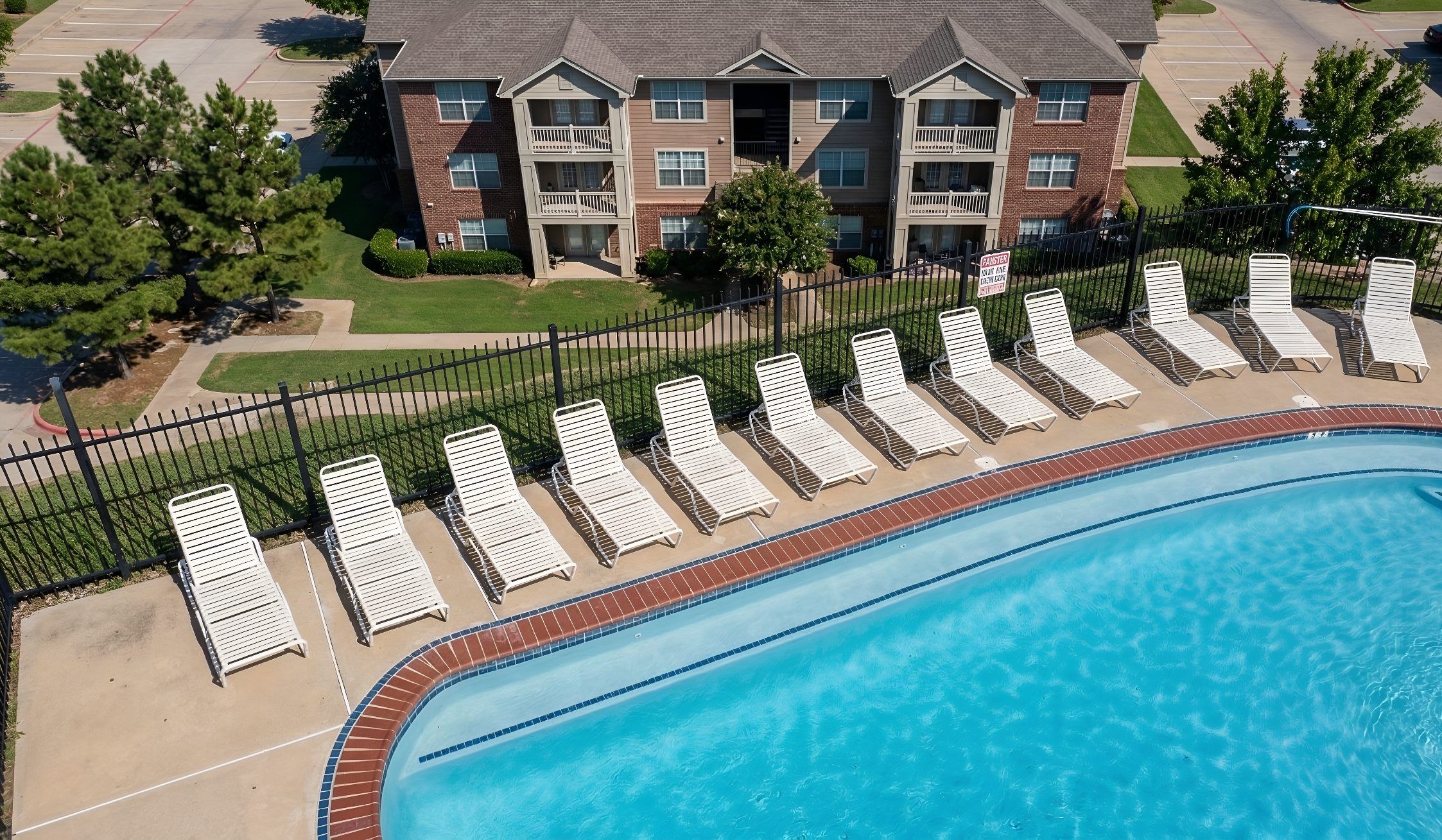 White lounge chairs line a poolside with an apartment building and trees in the background.